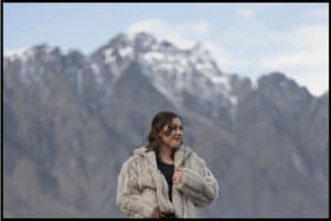 Young woman poses in a fur jacket in front of the snow capped Remarkables mountain range.