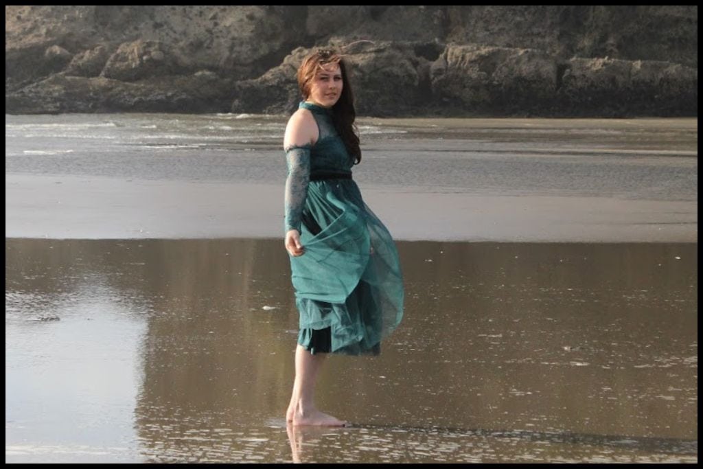 Young woman (author) in green tulle dress twirls on the reflective black sands of Piha Beach.