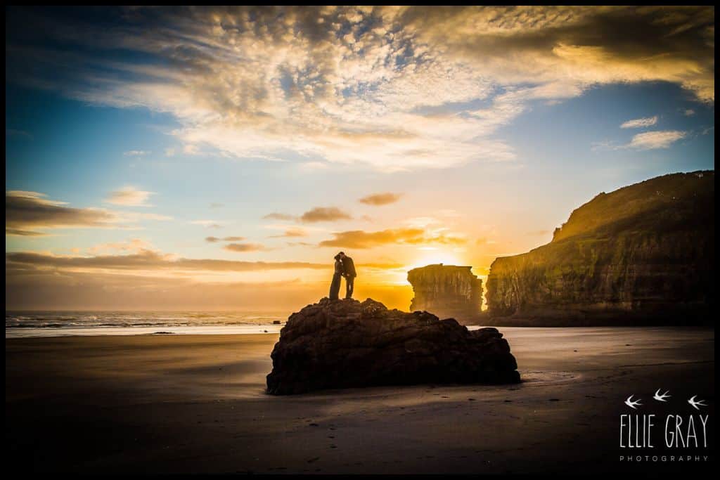 Silhouetted couple kiss at sunset atop the large boulder at Māori Bay.