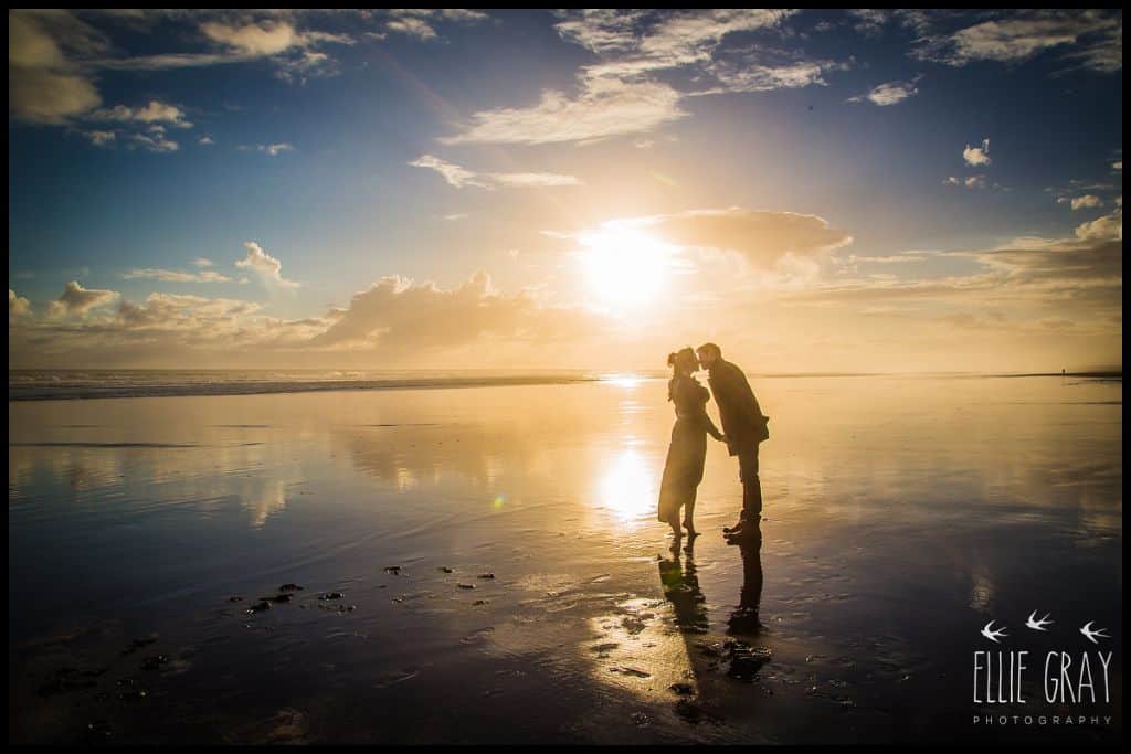 Silhouetted couple kiss on Muriwai Beach at sunset. The wet sand is as reflective as glass.