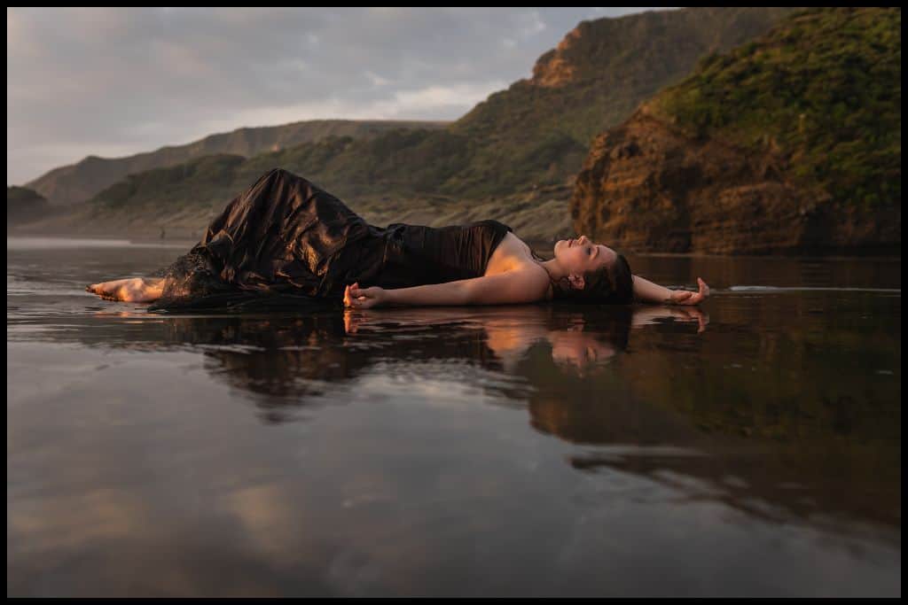 Young woman (author) in strapless gown lying on the wet sand, soaked, at Bethells Beach (Te Henga).