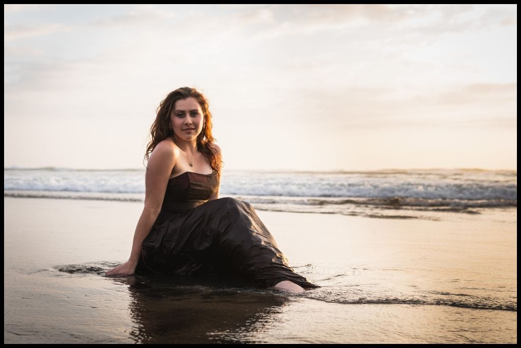 Young woman in strapless ball gown sits in the shallows at Bethells Beach.