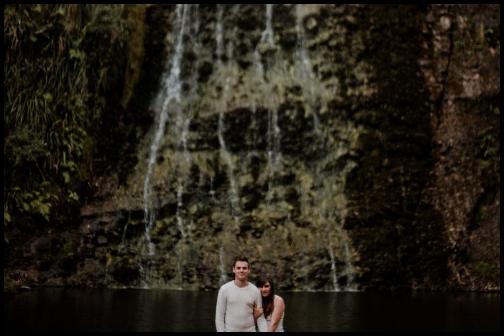 Young couple gaze at the camera with Karekare Falls in the background.