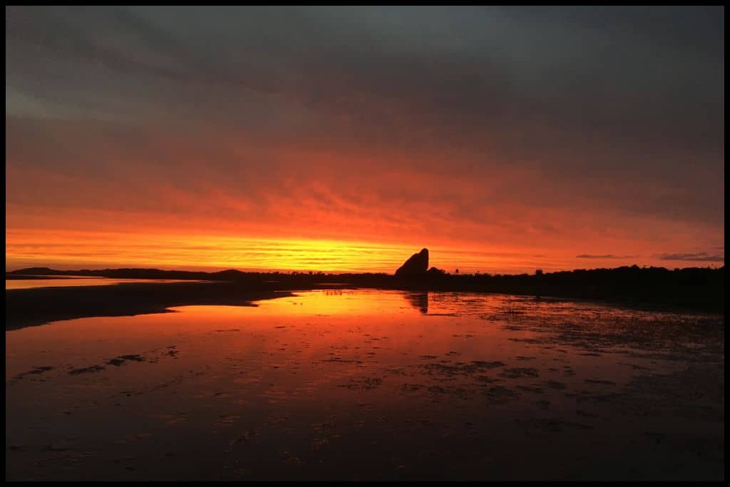 A fiery sunset captured from Whātipu Beach with Te Toka-Tapu-a-Kupe (Ninepin Rock) silhouetted against the sky.