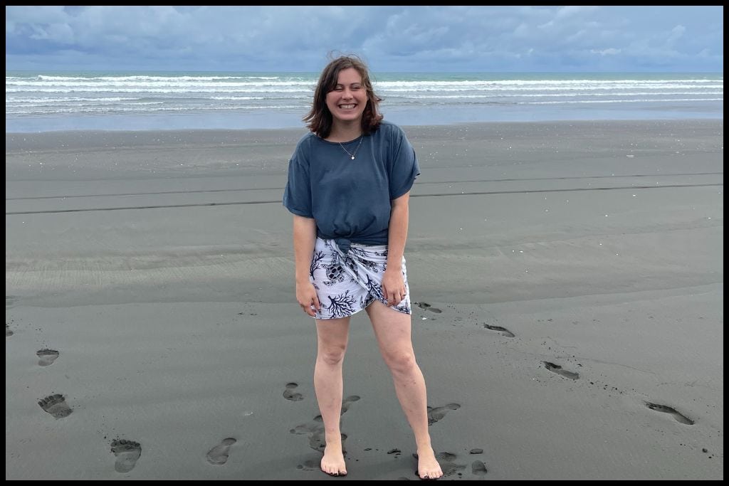 Young woman laughs at something behind the camera, with the black sands and waves of Kariotahi Beach in the background.