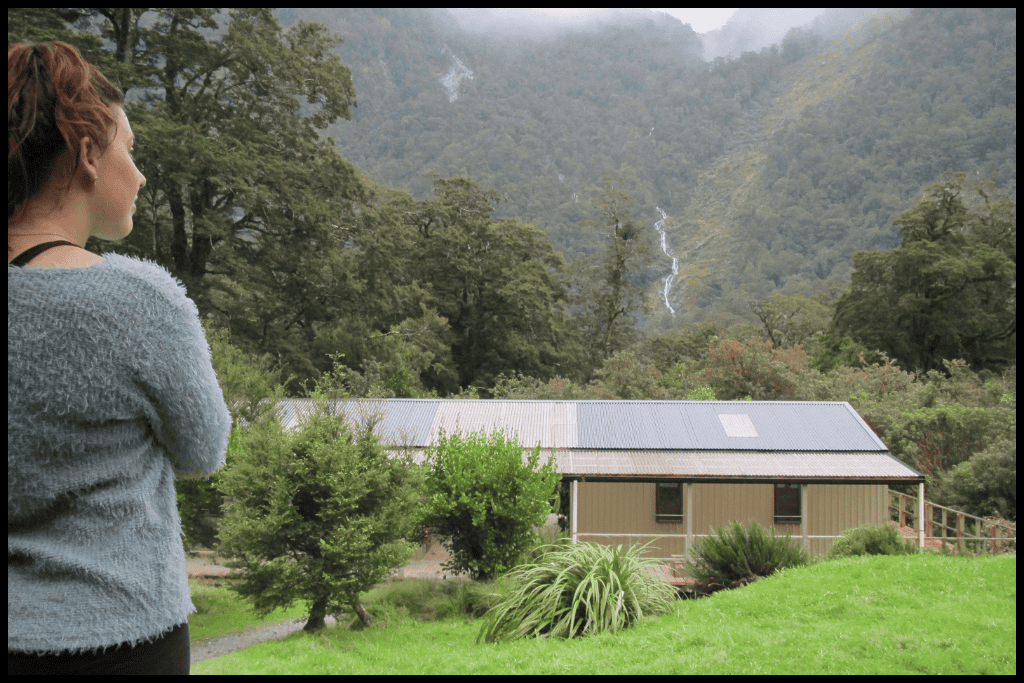 Author looks out to a distant waterfall with a Department of Conservation hut in the foreground.