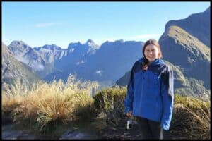 Author stands in blue raincoat atop the MacKinnon Saddle of the Milford Track with yellow tussock in the foreground and the Darren Mountain Range in the background.