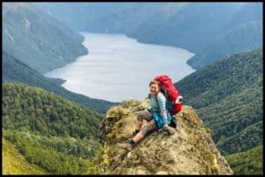 Young woman with a hiking pack on sits on a rocky outcrop overlooking the southern fjord of Lake Te Anau.