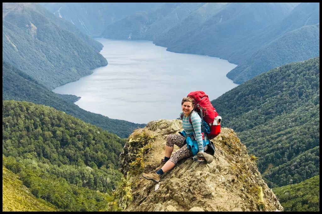 Young woman with a hiking pack on sits on a rocky outcrop overlooking the southern fjord of Lake Te Anau.