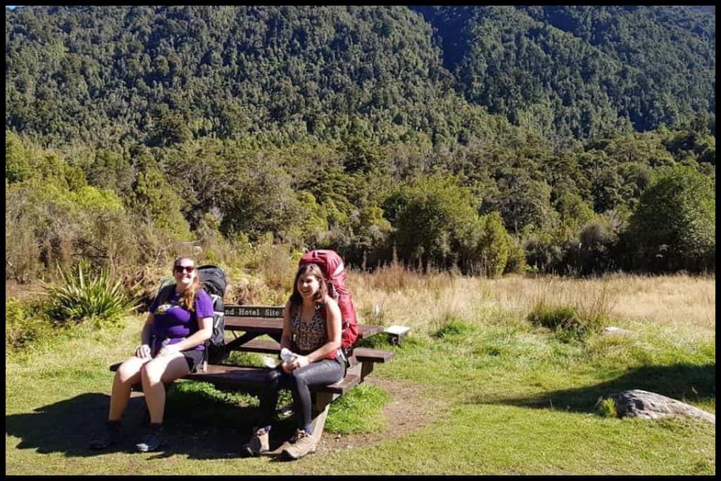 Two young women with their hiking packs on take a break on a picnic table by the side of the Paparoa Track.
