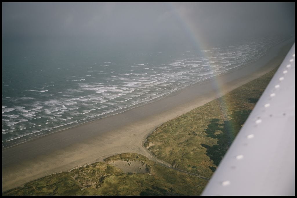 An aerial shot taken out of the plane window shows a rainbow refracted in the sky with the Stewart Island coastline below.