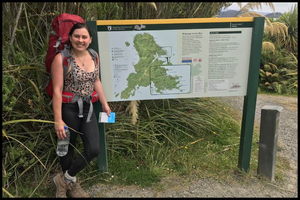 A young woman (the author) poses in her hiking gear next to the Lee Bay Road trailhead sign of the Rakiura Track.