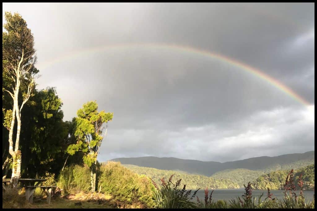 A rainbow stretches across a dark grey sky above the North Arm Hut, Stewart Island.