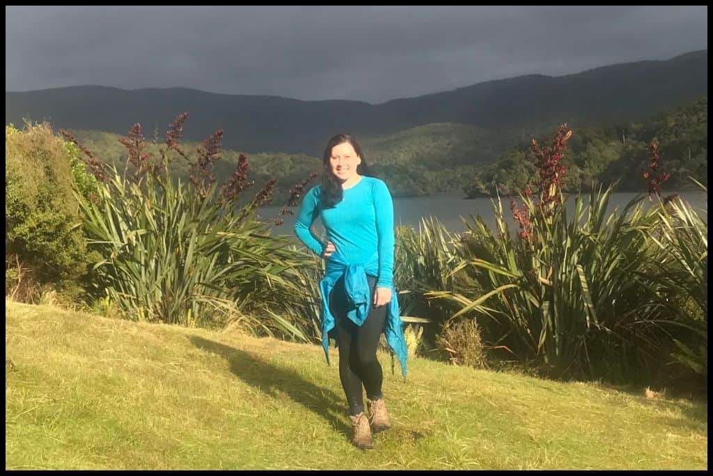 A young woman (the author) poses in her hiking gear in front of the North Arm Hut with a dark stormy sky behind her.