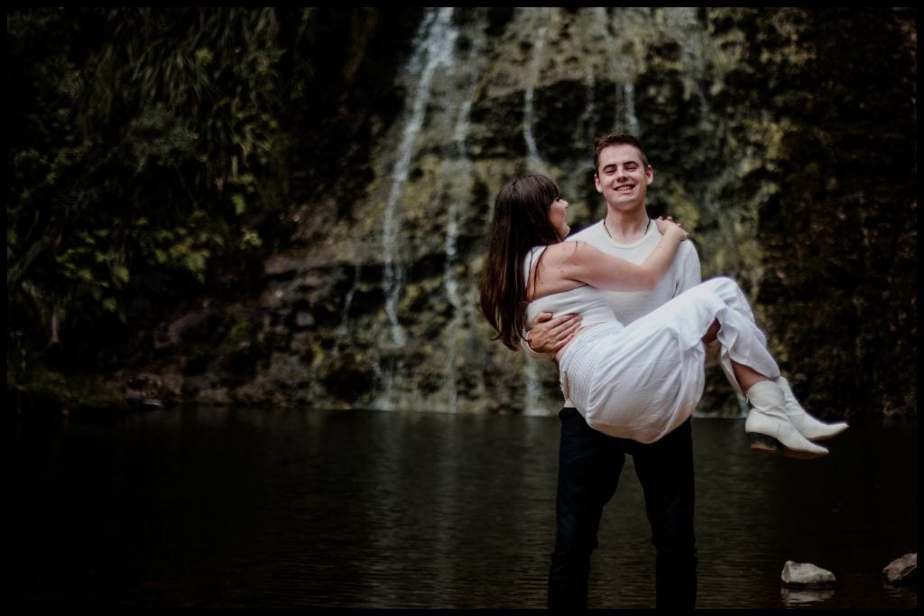 Young male scoops up his young wife in front of Karekare Falls.