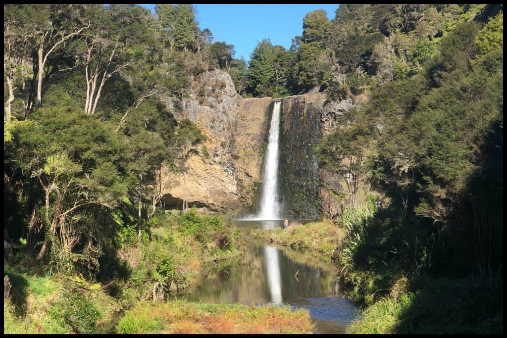Photo shows Hunua Falls under a blue sky, reflected in the creek in the foreground.
