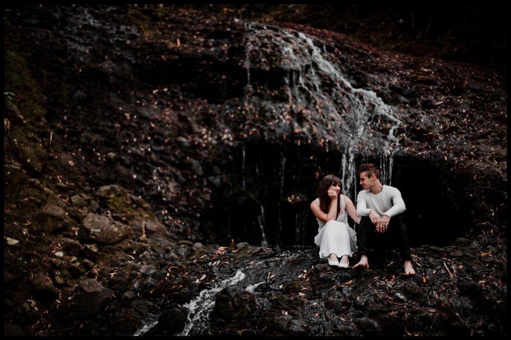Young couple in white sit perched against the black rocks with Opal Pool Stream Cascade trickling behind and beside them.