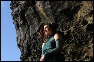 Young woman poses at black sand Piha Beach in a flowy dark green dress.