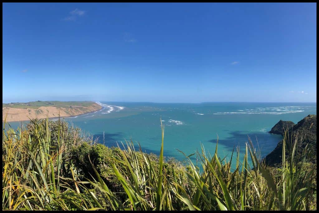 Turquoise coloured ocean, framed by the Āwhitu Peninsula across the Manukau Harbour, and the Signal House summit visible on the right.