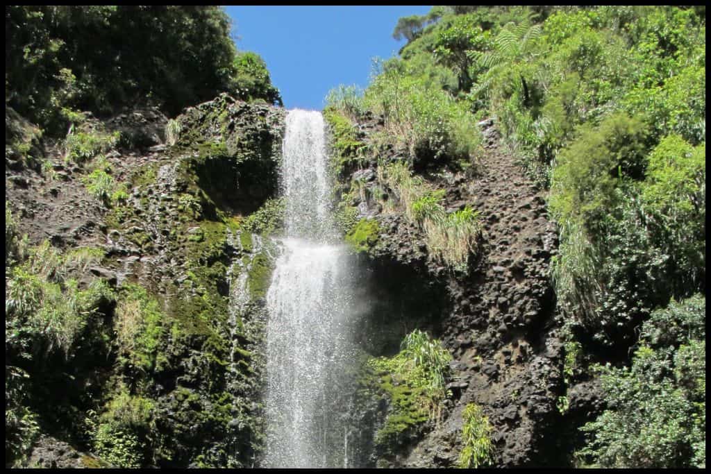 The top two tiers of Kitekite Falls cascade down over the rocks under a blue sky.