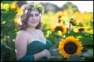 Young woman in a strapless green gown and sunflower crown cradles her baby bump behind the leaves of a tall sunflower.