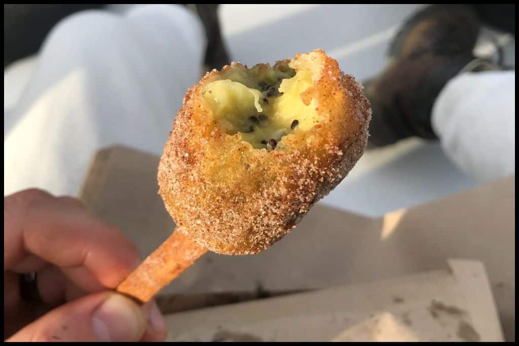 A close up shot shows a deep fried kiwifruit on a popsicle stick.
