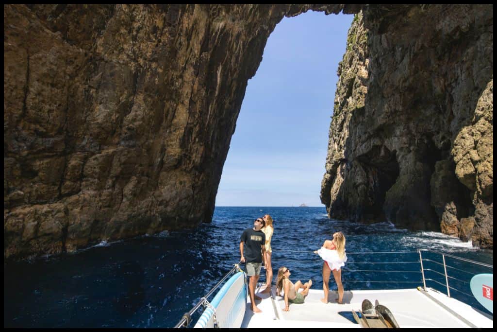 Photo shows some boat passengers on the deck out at sea under a hole in the rock.