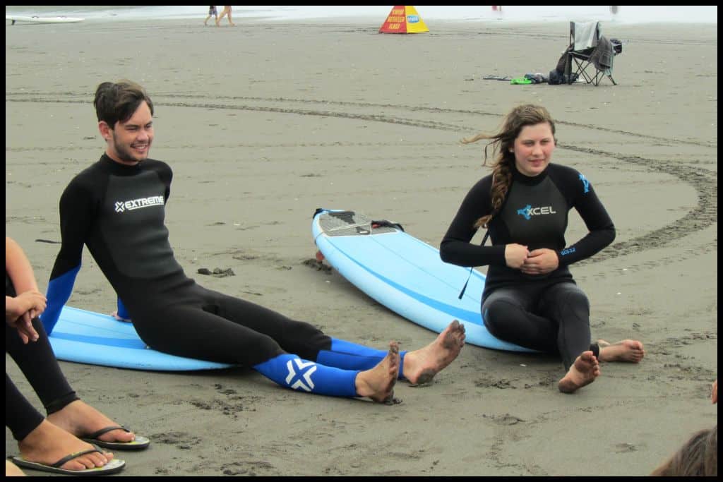Two young teens sit on their surfboards in wetsuits, picking up tips.