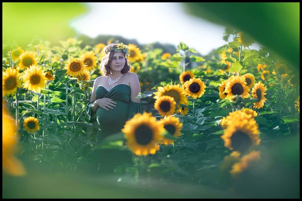 A pregnant woman in a green gown poses amongst dozens of tall sunflowers at a sunflower farm.