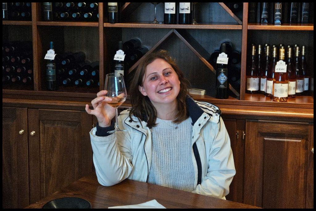 Young woman smiles for the camera, wine in hand, in a cellar door tasting room with an array of wine bottles behind her.