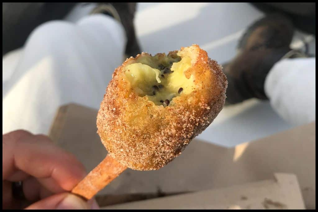 A close up shows a deep fried kiwifruit coated in sugar and skewered on a popsicle stick.