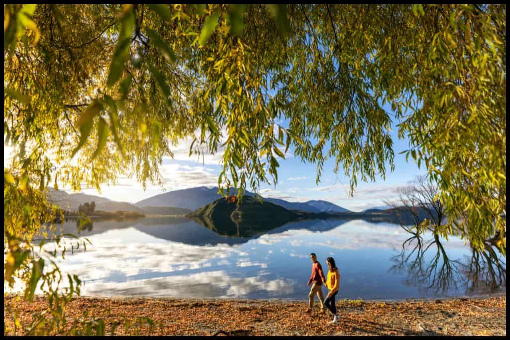 Two walkers stroll by Lake Wanaka. The photo is framed by overhanging poplar tree branches.