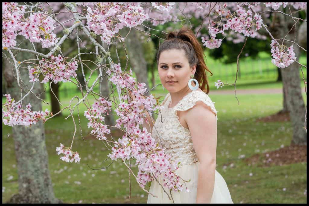 Young woman in cream dress poses amongst the cherry blossom trees.