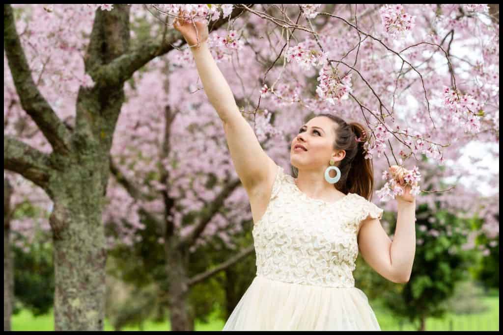 Young woman in cream dress poses below the cherry blossom trees.