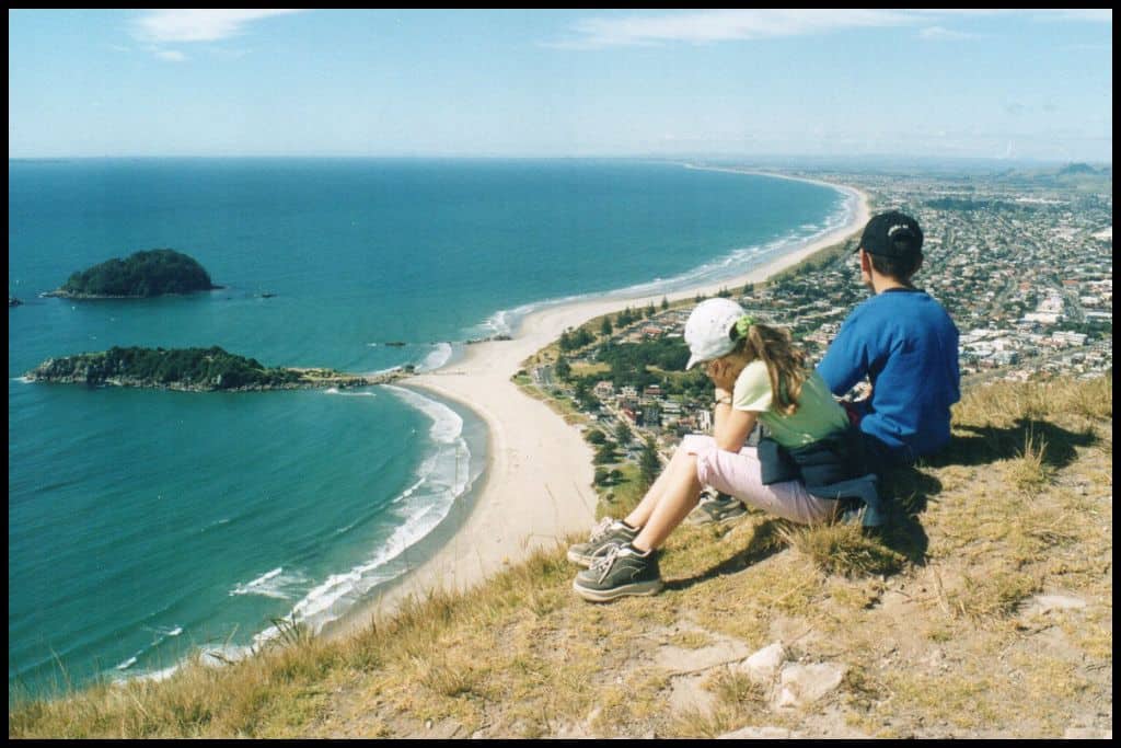 Two children sit on the summit of Mount Maunganui looking down to the beach below.