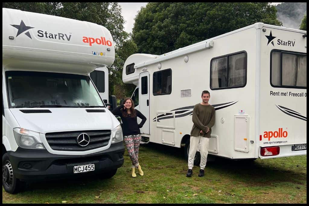 A young couple leans against a pair of motorhomes, parked up at a scenic reserve.