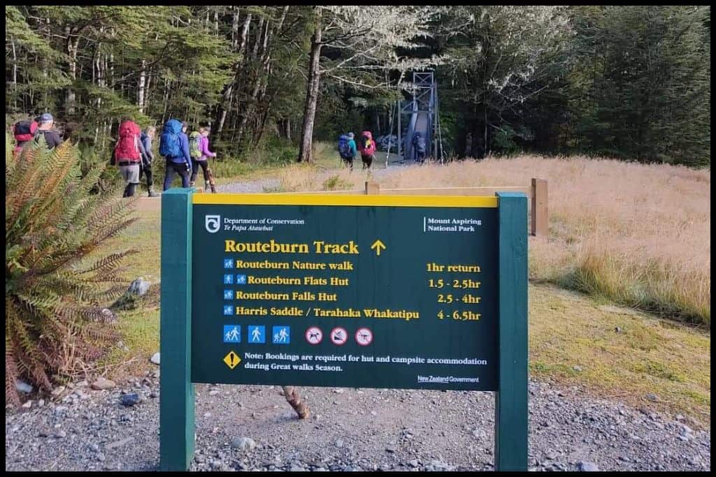 A closeup of the Routeburn Track sign, taken from the Routeburn Shelter end of the track, with the first swingbridge and a group of hikers in the background.