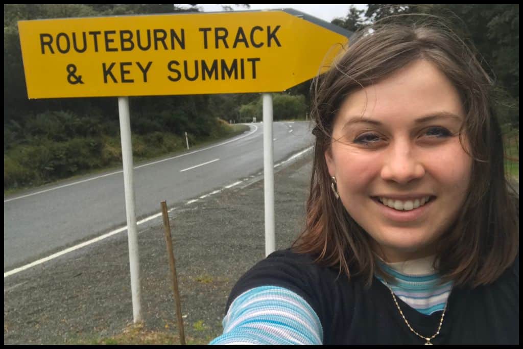 A young woman smile for selfie in front of the Routeburn Track sign at The Divide end of the track.