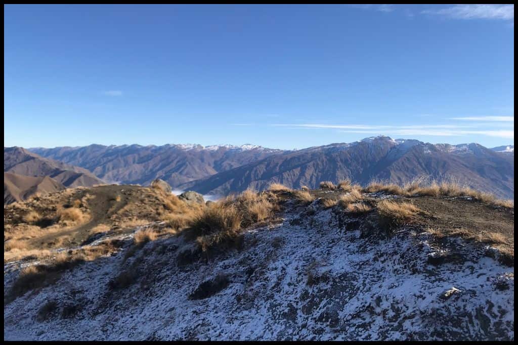 View taken from atop Roys Peak shows snow in the tussock in the foreground and a dusting of snow on the mountains in the background.