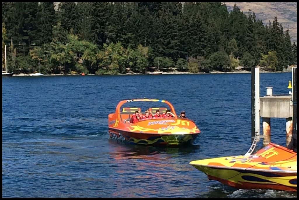 A orange jet boat cruises back into the wharf across a dark blue Lake Wakatipu.