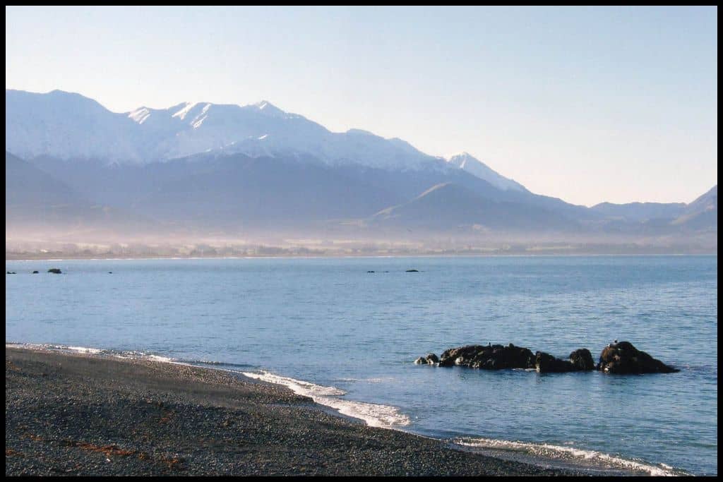 Snow capped mountains overlook black pebbled Kaikoura Beach in early morning sunshine.