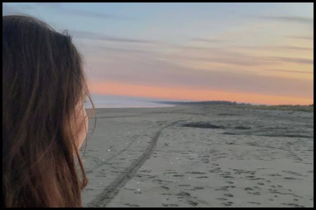 Side profile of a woman looking over her shoulder at the sandy beach and setting sun.