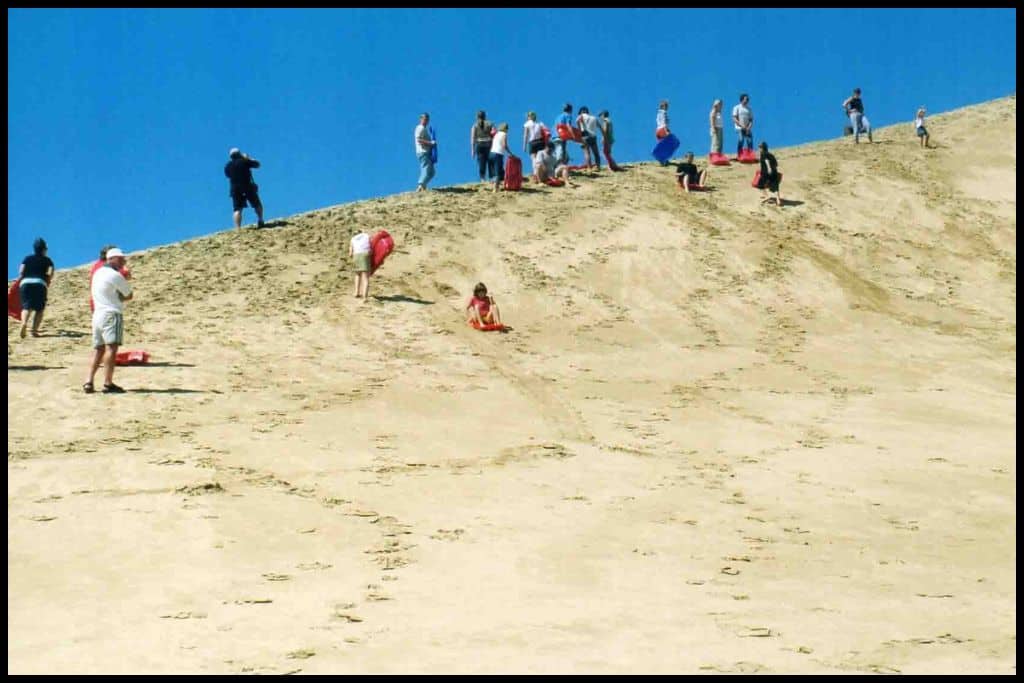 A group of tourists stand atop the Te Paki Sanddunes as a few make their way down on toboggans.