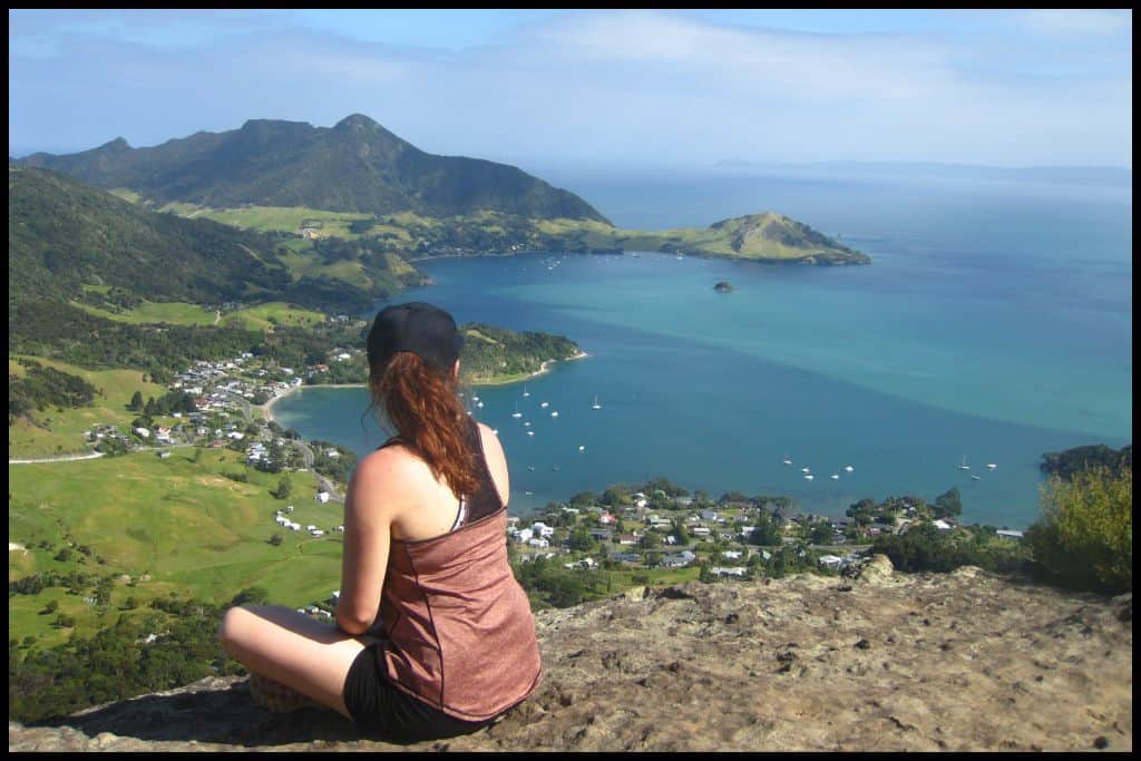 A young female sits crosslegged on a rocky outcrop at the summit of Mount Manaia with Whangārei Harbour and Heads below.
