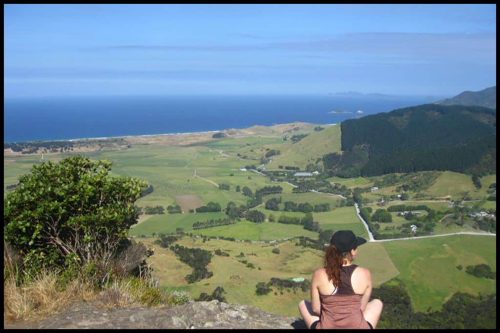 Young female sits crosslegged on a rocky outcrop at the top of Mount Manaia, with a very straight, deep blue stretch of coastline (Ocean Beach and Bream Head) in front of her, separated only by rolling green paddocks.