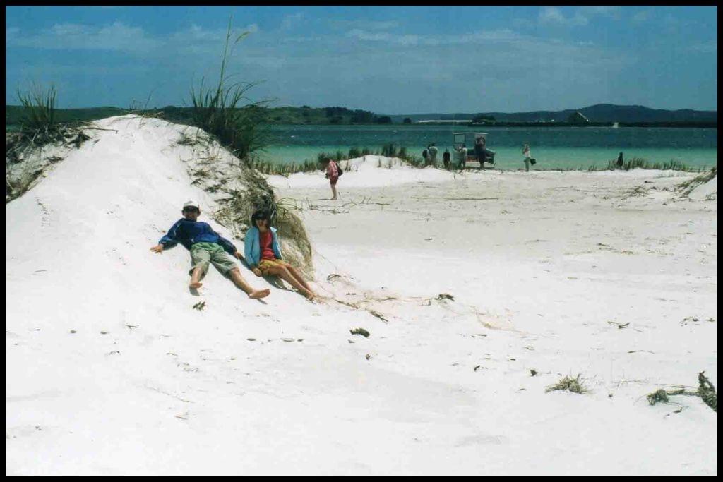Two children lean up against a mound of white silica sand with part of the clear, aqua coloured Parengarenga Harbour visible in the background.