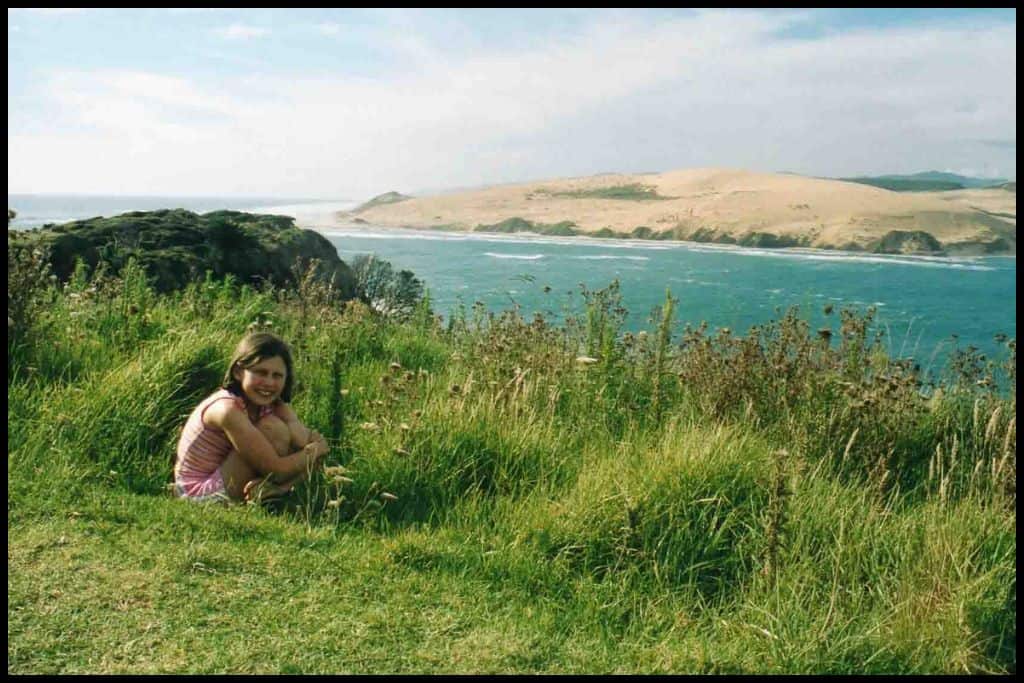 Young girl crouches in the grass with the sand dunes marking the entrance to the Hokianga Harbour in the background.