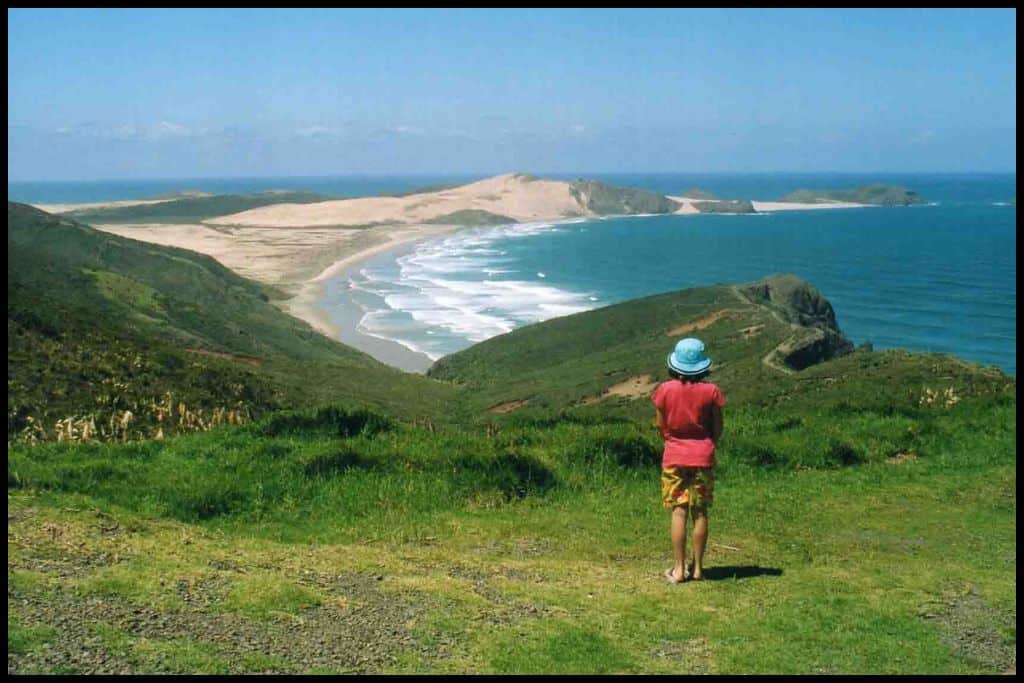 Young girl stands looking out to the sand dunes and surf at Te Werahi Beach.