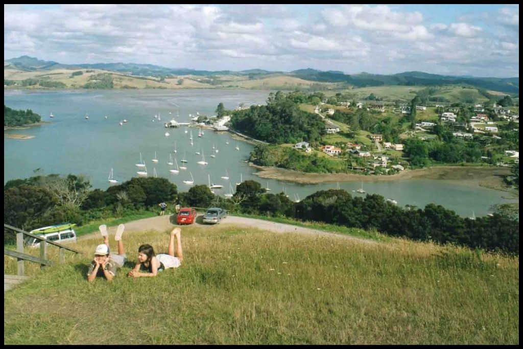 Two kids lie in the grass up the top of a grassy hill overlooking the Mangōnui Harbour.