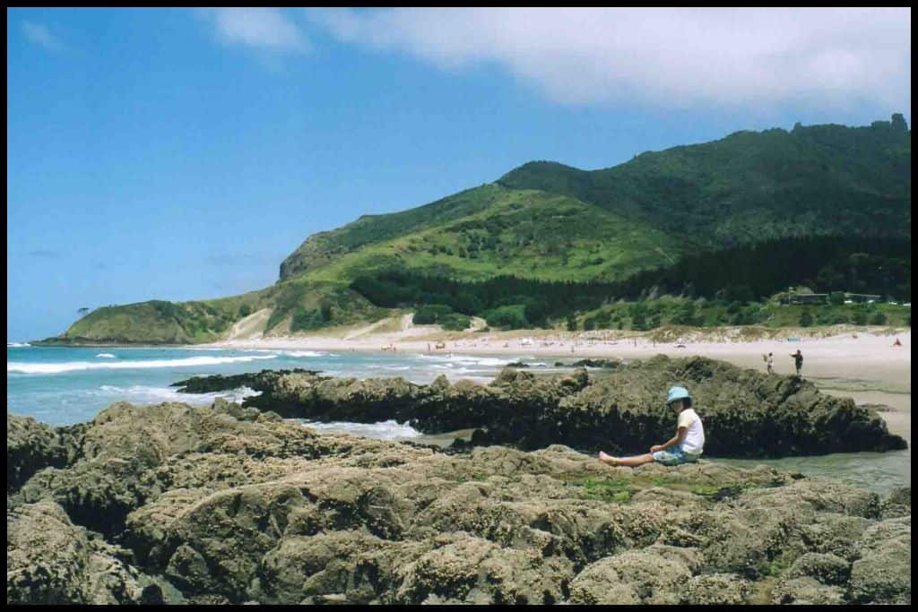 A young girl sits on the rocks at the end of Ocean Beach, Breams Head visible in the background.